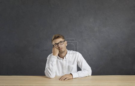 Photo for Portrait of young pensive male employee sitting at table and bored, tired and need relax day. Caucasian man sitting at table on gray background and looking up at copy space above his head. - Royalty Free Image