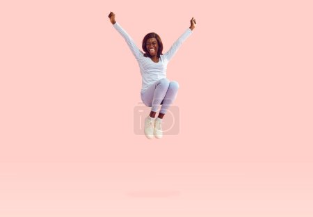Happy young energetic African American woman in casual blue long sleeve shirt, skinny jeans and white sneakers jumping on trampoline and having fun high in air isolated on solid light pink background