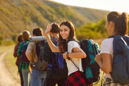 Téléchargez les photos : Joyeux sourire jolie fille brune agitant la main regardant joyeusement la caméra tout en trekking avec sac à dos lors d'une visite à pied des montagnes avec un groupe de ses jeunes amis. Concept de randonnée. - en image libre de droit