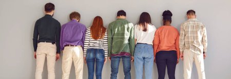 Back portrait of multiethnic diverse group of a young people friends or colleagues wearing casual clothes standing in a row on gray wall background. Rear view of men and women in a line. Banner.