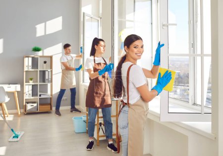 Cheerful caucasian woman, part of a dedicated cleaning staff team, meticulously wipes windows during a home or office service, demonstrating professionalism and teamwork in household chores.
