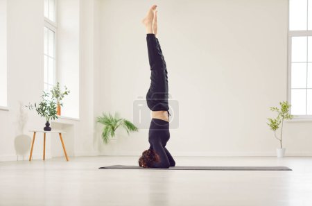 Photo for Young attractive woman practicing yoga, standing in salamba sirsasana exercise, headstand pose, working out wearing black sportswear in white studio, home, meditating alone for health balance - Royalty Free Image