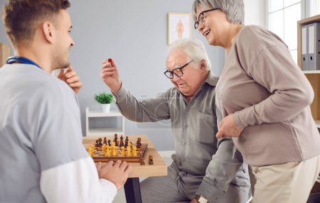 Téléchargez les photos : Jeune homme infirmier jouant au jeu d'échecs avec un heureux sourire joyeux couple âgé assis à la table dans une clinique médicale. Soutien aux aidants naturels et concept de loisirs pour la maison de retraite. - en image libre de droit