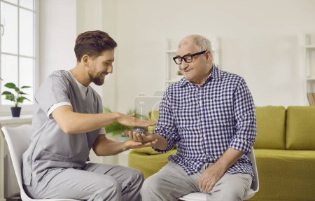 Foto de Retrato de un joven fisioterapeuta amigable sonriente, enfermera o cuidador mostrando a un hombre mayor de pelo gris un ejercicio con una bola de pinchos para el masaje de manos sentado en casa en la jubilación. - Imagen libre de derechos