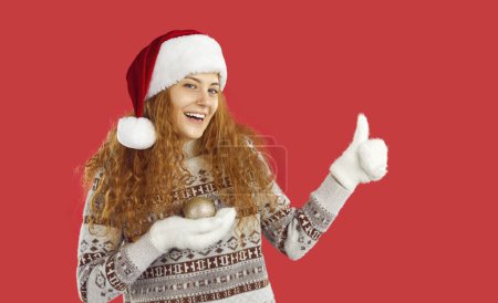 Photo for Girl is advertising Christmas sale or special offer. Portrait of cheerful cute young woman holding Christmas tree toy in her hand and showing thumb up. Woman in Santa hat isolated on red background. - Royalty Free Image
