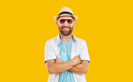 Porträt eines selbstbewusst lächelnden jungen bärtigen, gutaussehenden Mannes mit verschränkten Armen, der lässige Sommerkleidung mit Hut und Sonnenbrille auf gelbem Hintergrund im Studio trägt.