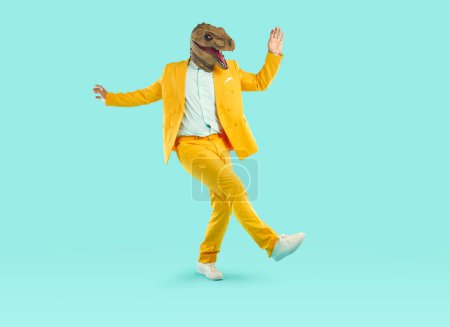 Téléchargez les photos : Jeune homme énergique dansant portant un masque de tête de dinosaure. pleine taille photo de bizarre excentrique gars portant élégant costume jaune s'amuser sur fond de studio isolé - en image libre de droit