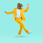 Energetic young man dancing wearing dinosaur head mask. Full size photo of freaky eccentric guy wearing stylish yellow suit having fun over isolated studio background