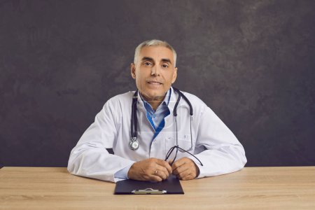 Photo for Portrait of smiling middle aged doctor in lab coat with stethoscope around his neck. Friendly caucasian male healthcare worker looking straight into camera while sitting at desk on gray background. - Royalty Free Image