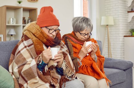 Téléchargez les photos : Couple âgé assis ensemble sur le canapé dans le salon à la maison, orné de vêtements d'hiver. Enveloppés dans une couverture réconfortante, ils bercent des tasses de thé fumantes, essayant de se réchauffer pendant l'hiver. - en image libre de droit