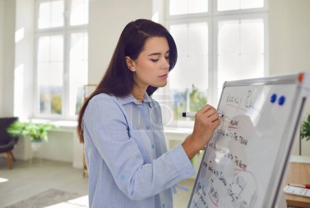 Vue latérale d'une jeune femme d'affaires debout devant le tableau blanc planificateur tâches de planification sur une semaine tenant marqueur en main. Travailleuse écrivant dans le calendrier calendrier avec rendez-vous plan quotidien.