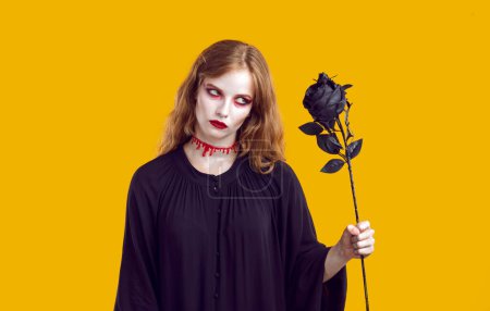 Téléchargez les photos : Portrait of mysterious scary dead woman with black rose in her hand on orange background. Witch woman in black dress and with blood on her neck looks at rose with terrible look. Halloween concept. - en image libre de droit
