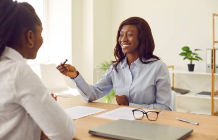 Foto de Dos jóvenes afroamericanas se reúnen en la oficina, negocian un acuerdo de negocios y firman un contrato. Mujer sonriente sentada en una mesa le da un bolígrafo y un documento en papel a su cliente de negocios - Imagen libre de derechos