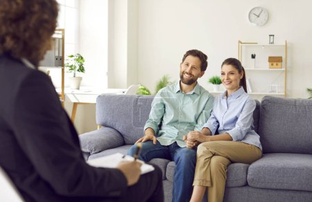 Photo for Happy smiling young couple sitting on the couch at counselors office, listening advices and smiling holding hands. Man and woman talking with male family psychologist during therapy session. - Royalty Free Image