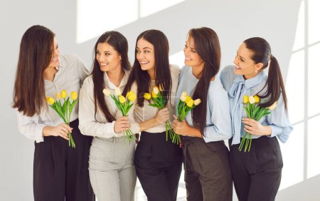 Photo for Group of beautiful happy smiling women with yellow flowers tulips bouquet standing in a line on a light gray background and talking celebrating March 8. International Womens Day and spring concept. - Royalty Free Image