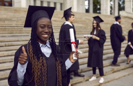 Foto de Joven feliz sonriente afroamericana estudiante femenina en un vestido de graduado universitario y diploma en sus manos. Graduada chica mostrando pulgar hacia arriba signo al aire libre con compañeros de clase en el fondo. - Imagen libre de derechos