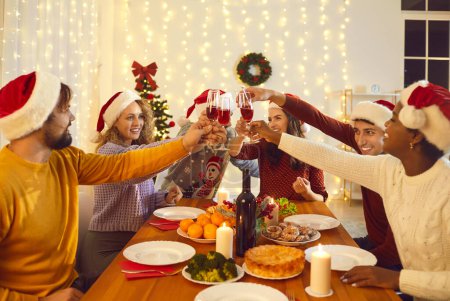 Foto de Grupo de alegres jóvenes amigos multirraciales celebran la Navidad en un ambiente acogedor en casa. La gente en Santa Claus sombreros tintineo vasos de vino tinto mientras se sienta en la mesa festiva durante la cena de Navidad. - Imagen libre de derechos