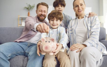 Photo for Smiling family, parents and children, is putting a coin into a piggy bank together at home sofa. Act of saving money as a family and the joy of working towards a common financial goal. - Royalty Free Image