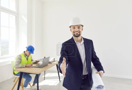 Portrait of happy young smiling man architect extending his hand to camera to make a deal standing on building site with documents in hands and with foreman working at desk in background.