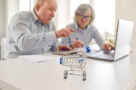 Primer plano de un carrito de compras de juguetes con una pareja de ancianos, trabajando en una computadora portátil en casa. Gestión conjunta de pensiones, inversiones o compras en línea, ilustrando las actividades y responsabilidades compartidas.