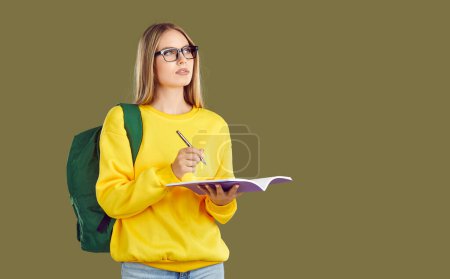 Ernsthaft denkende blonde Studentin in Brille und gelbem Sweatshirt mit Rucksack macht sich Notizen in Textbüchern auf khakifarbenem Hintergrund und schaut auf den Kopierraum. Bildungsbanner für Werbekonzept.