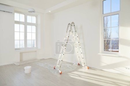 Photo for Step ladder, paint bucket and piece of floor protective film in corner of modern living room with freshly painted white walls, plastic windows and no people. Home renovation, apartment renewal concept - Royalty Free Image