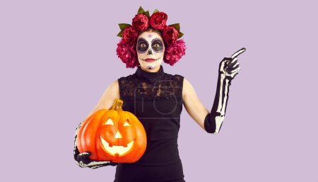 Téléchargez les photos : Fille avec du maquillage de crâne de sucre pointant avec son doigt sur le côté. Jeune femme portant une couronne de fleurs, une robe noire et des gants squelette tenant une citrouille effrayante et des gestes. Concept de fête d'Halloween - en image libre de droit