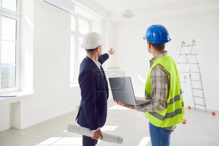 Young foreman discussing with professional male architect design project or repair of new apartment on a laptop standing back in empty room. Meeting with builder about interior decoration.