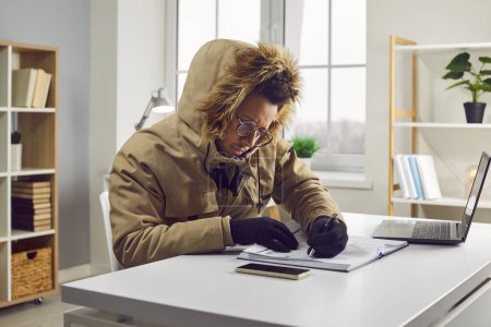 Portrait of young indian business man in winter coat with hood and gloves working in the cold at home on his laptop computer at the desk trying to keep warm and writing. Heating problems concept.