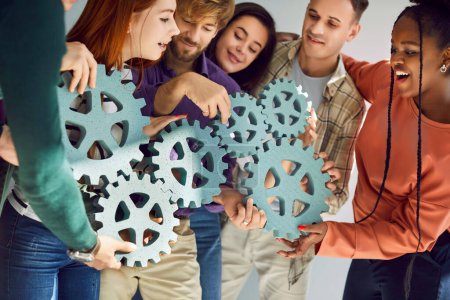 Team of diverse people connecting mechanical cogs. Group of smiling male and female teammates holding symbolic gear wheels in hands and joining them all in one chain. Cooperation and teamwork concept
