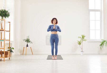 Photo for Yogi young female instructor in sportswear standing with namaste gesture, enjoying meditation practice, starting exercising for life balance, working out calm in studio, home among green houseplant - Royalty Free Image