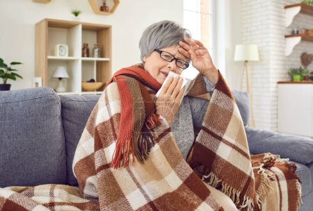 Téléchargez les photos : Sick senior woman with runny nose wrapped in a blanket sitting on sofa at home. Elderly person with seasonal flu sneezing in napkin, feel unhealthy with influenza and touching her forehead with hand. - en image libre de droit