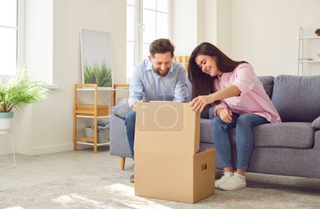 Photo for Happy young joyful couple unpacking parcel at home, sitting in the living room on sofa and smiling looking into open cardboard box. Delivery, online shopping, receive the parcel concept. - Royalty Free Image