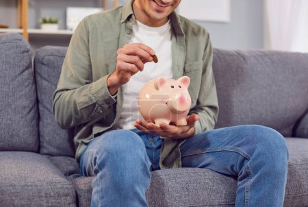 Man sits on a couch at home, putting a coin into a piggy bank. He is happy and focused, planning money budget and ensuring financial wealth for the future, demonstrating responsible financial habits.
