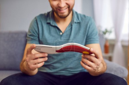 Positive man is sitting on the sofa, inspecting an orthopedic insole. Hands holding the insole, emphasizing health and medical care for feet. Perfect for orthopedics related concepts.