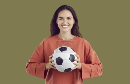 Soccer fan happy smiling brunette woman holding football ball in hand on khaki background and looking at camera. She is wearing orange sweatshirt. Celebrating victory, winner, soccer, football game.