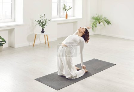 Jeune femme attrayante sportive yogi pratiquant le yoga, assise dans l'exercice Ustrasana, pose de chameau, faisant de l'exercice, portant des vêtements de sport blancs, fond intérieur de la maison, près des plantes en pot