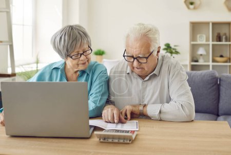 Senior couple sitting at the table with laptop looking at the bills calculating finances or taxes at home. Elderly retired man and woman counting income and profit. Pension calculation concept.