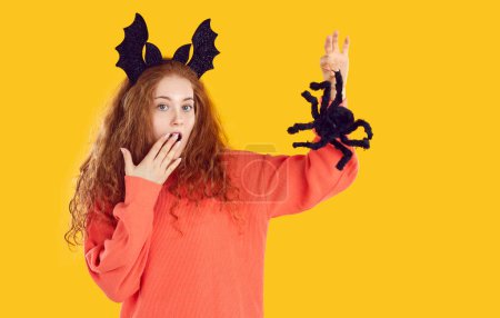 Celebración de Halloween. Mujer divertida imitando mirada asustada mientras sostiene espeluznante araña decorativa de Halloween. Mujer rizada pelirroja con aro de Halloween pone su mano en la boca sobre fondo naranja vivo.