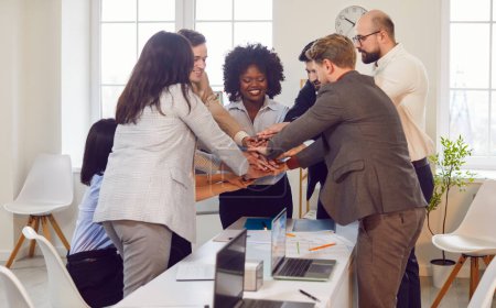 Group of business people standing in a circle in an office, stacking hands together. Gesture signifies unity, teamwork, team collaboration, collective effort and commitment to achieving common goals.