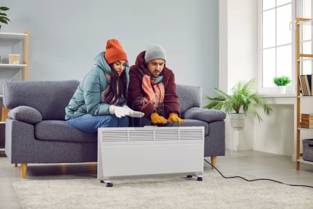 Full length photo of a young frozen couple sitting on the sofa in the living room in winter outerwear and hats at home and trying to warm their hands on electric heater. Heating problems concept.
