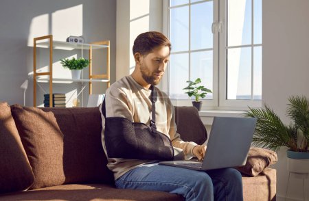 Junger Mann mit gebrochenem Arm in einer medizinischen Armschlinge, der von zu Hause aus arbeitet, auf dem Sofa sitzt, seinen modernen Laptop benutzt und mit der linken Hand auf der Tastatur tippt