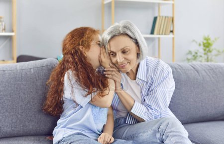 Happy granddaughter whispers a secret or gossip into grandmother ear. This intimate family moment captures the joy of sharing hearsay and talking together, highlighting close bond.