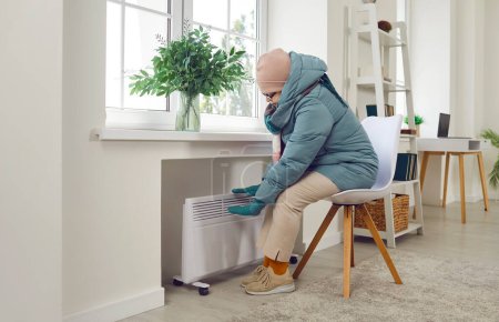 Aînée triste femme âgée assise sur une chaise en vêtements d'hiver dans le salon réchauffer ses mains sur le chauffage électrique à la maison. Personne mûre gelant à la maison. Problèmes de chauffage concept.