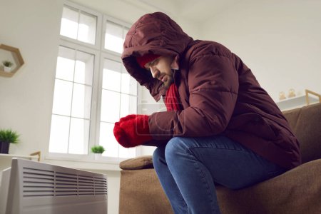Portrait of a young disappointed worried frozen man sitting on sofa in winter outerwear in hood at home and trying to warm his hands on modern electric heater. Heating problems concept.