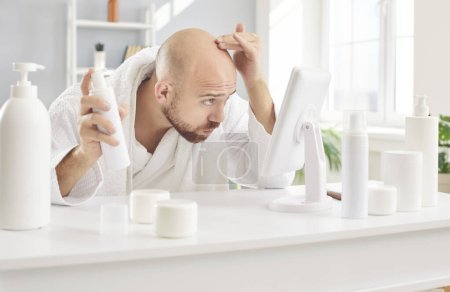 Concerned balding young man in white bathrobe sitting at table with sprays and shampoos for damaged roots and premature hair loss issue treatment, looking in mirror, and rubbing his top and forehead