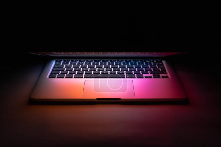Téléchargez les photos : Un ordinateur portable à moitié fermé dans le noir avec une lueur colorée - en image libre de droit