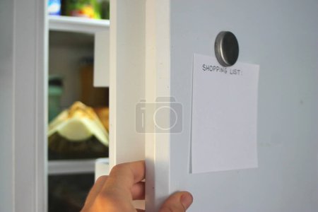Einkaufsliste mit Magnet am offenen weißen Kühlschrank