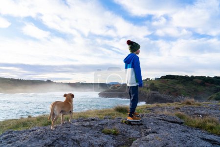 Boy with his dog on a cliff looking at the landscape with the sea in the background