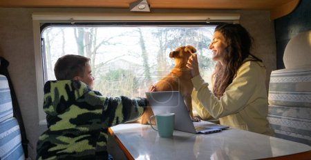 Téléchargez les photos : Mère célibataire voyageant avec son fils et son chien dans un camping-car et travaillant sur l'ordinateur portable - en image libre de droit